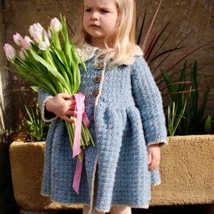 May include: A young girl wearing a light blue crocheted coat with a white collar and a pink ribbon. She is holding a bouquet of pink tulips.