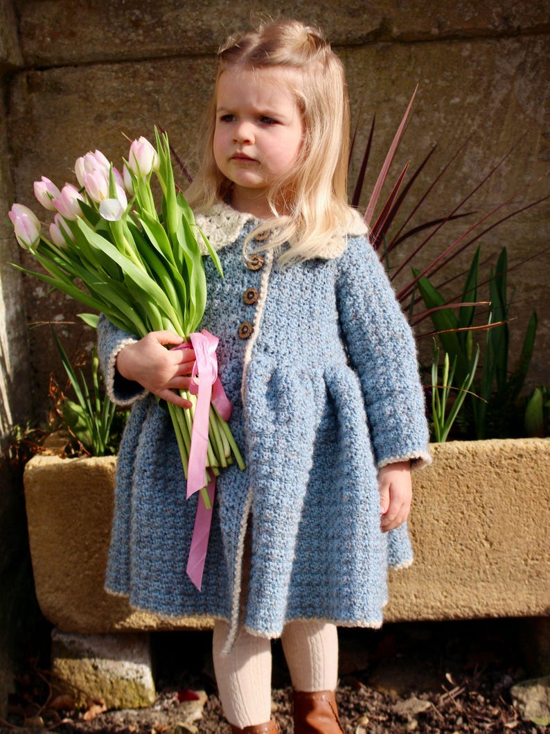 May include: A young girl wearing a light blue crocheted coat with a white collar and a pink ribbon. She is holding a bouquet of pink tulips.