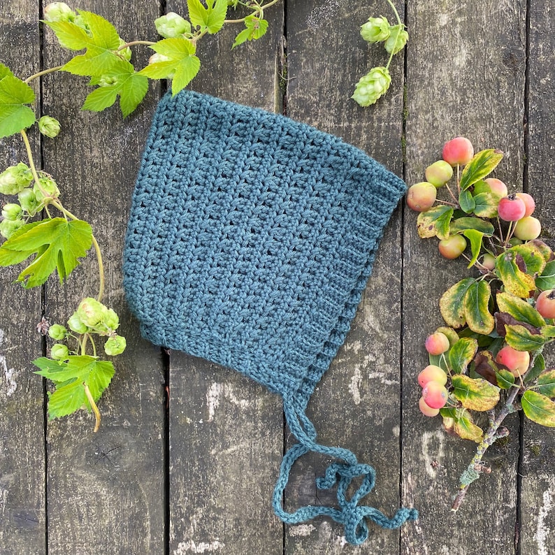 Puede incluir: Un gorro de beb&eacute; de ganchillo azul verdoso con un patr&oacute;n de punto sencillo. El gorro tiene lazos que se pueden atar debajo de la barbilla.