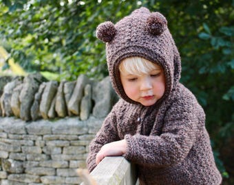 May include: A dark brown crocheted hooded jacket featuring two small pom-poms on top of the hood to resemble ears. The garment is crafted with a textured stitch pattern and includes a single button closure at the front neckline area.