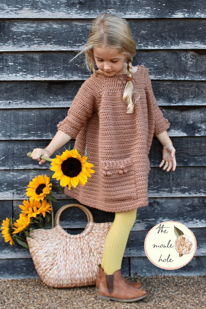 May include: A young girl wearing a brown crocheted dress with a pocket and yellow tights. She is holding a bouquet of sunflowers and a woven tote bag. The image includes the text "The moule hole".