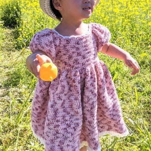 Op de afbeelding: Een jong meisje draagt een roze en witte gehaakte jurk met een witte gehaakte kantversiering. Ze draagt een strohoed en houdt een geel speelgoed vast. Het meisje staat in een veld met gele bloemen.