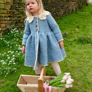 May include: A young girl wearing a light blue crocheted coat with a white lace collar and a wooden carrying basket with pink ribbon and pink and white tulips.
