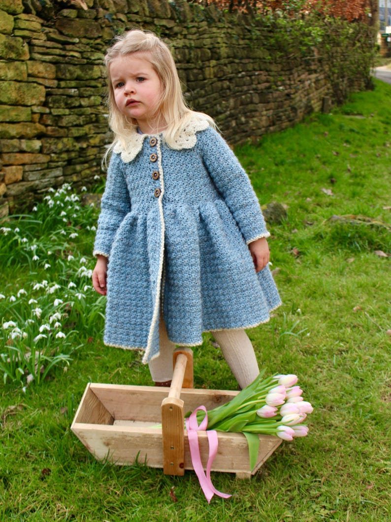 May include: A young girl wearing a light blue crocheted coat with a white lace collar and a wooden carrying basket with pink ribbon and pink and white tulips.