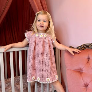 Può includere: Una bambina che indossa un vestito rosa all'uncinetto con dettagli di margherite bianche. Il vestito ha maniche corte e una scollatura rotonda. La bambina è in piedi davanti a un lettino e una poltrona rosa.