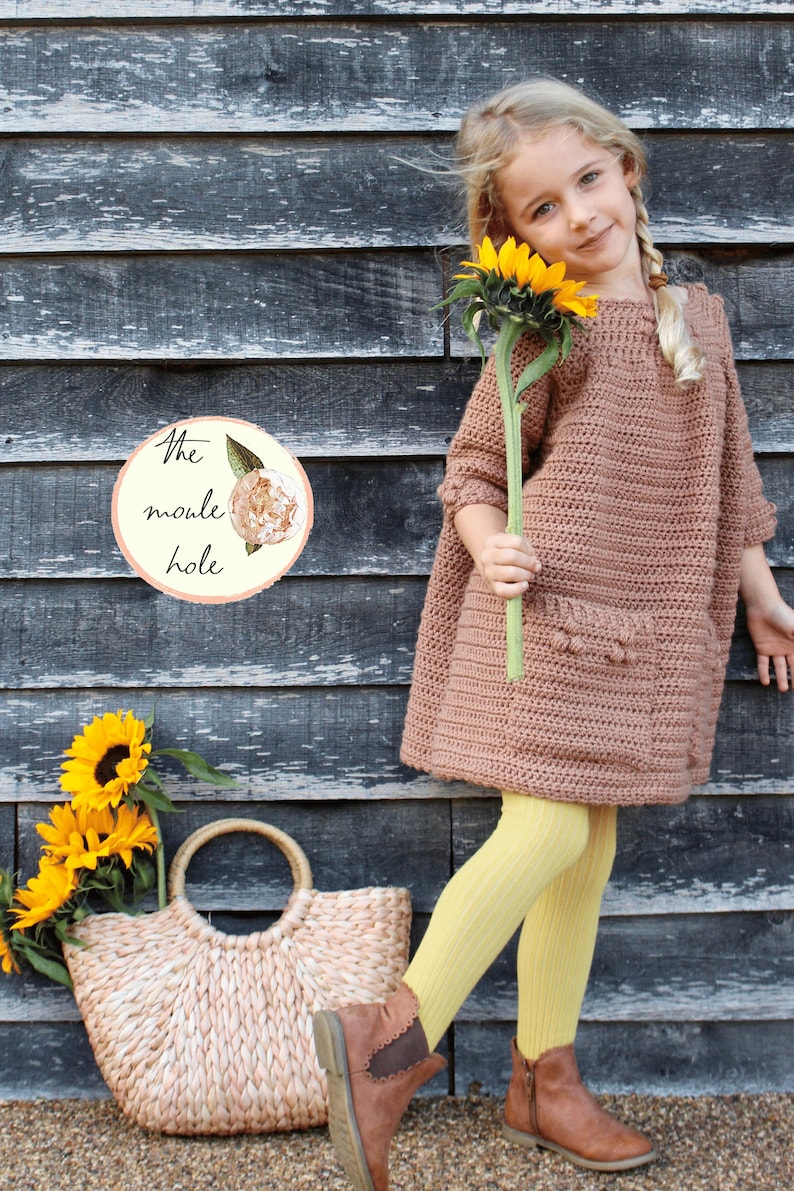May include: A young girl wearing a brown crocheted dress, yellow tights, and brown boots holds a sunflower. She is standing in front of a wooden wall with a sign that reads "The moule hole".