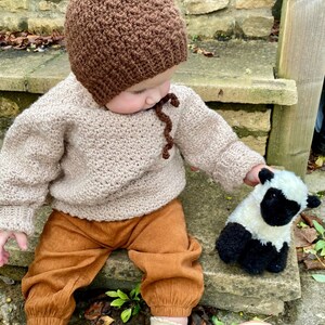 May include: A baby wearing a brown crocheted hat with a pointed top and a beige crocheted sweater. The baby is sitting on a stone step and holding a white and black stuffed sheep.