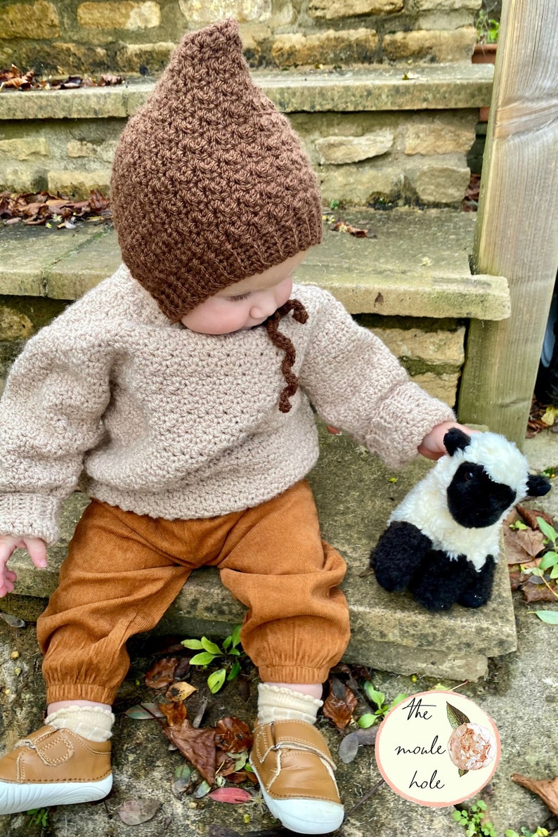 May include: A baby wearing a brown crocheted hat with a pointed top and a beige crocheted sweater. The baby is sitting on a stone step and holding a white and black stuffed sheep.