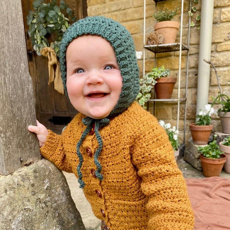 Puede incluir: Un beb&eacute; sonriente que lleva una boina de ganchillo verde y un c&aacute;rdigan de ganchillo marr&oacute;n. El beb&eacute; est&aacute; de pie cerca de un poste de madera y una piedra.