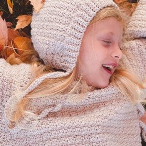 May include: A young girl wearing a light pink crocheted sweater and matching hooded hat, lying on a bed of autumn leaves.
