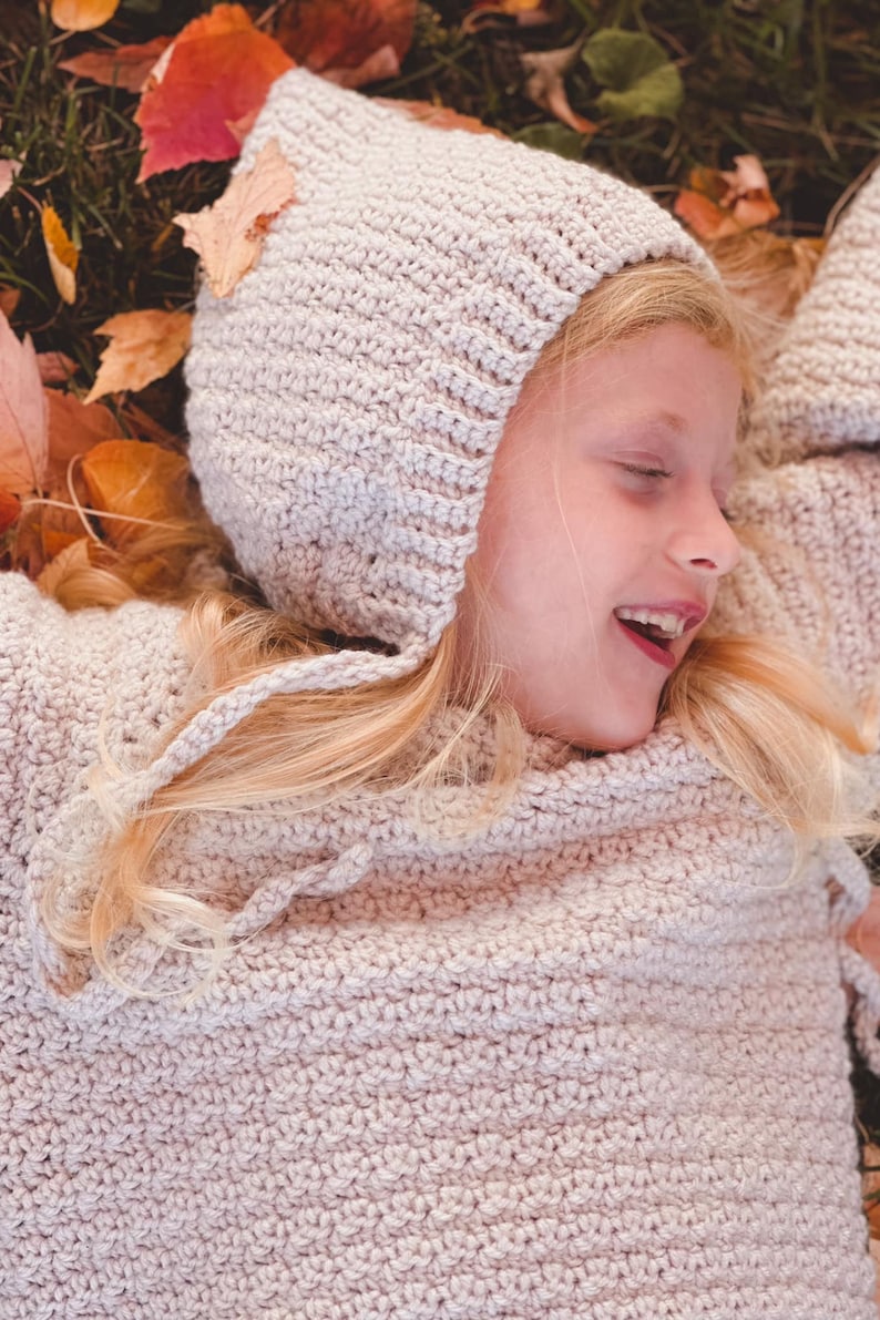 May include: A young girl wearing a light pink crocheted sweater and matching hooded hat, lying on a bed of autumn leaves.