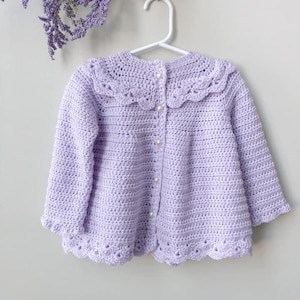 May include: A lavender crocheted cardigan with a scalloped collar and pearl buttons. The cardigan has a delicate crocheted design and is perfect for a baby or toddler.