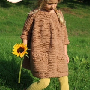 May include: A young girl wearing a brown crocheted dress with pockets and yellow tights. She is holding a sunflower and standing in a grassy field.