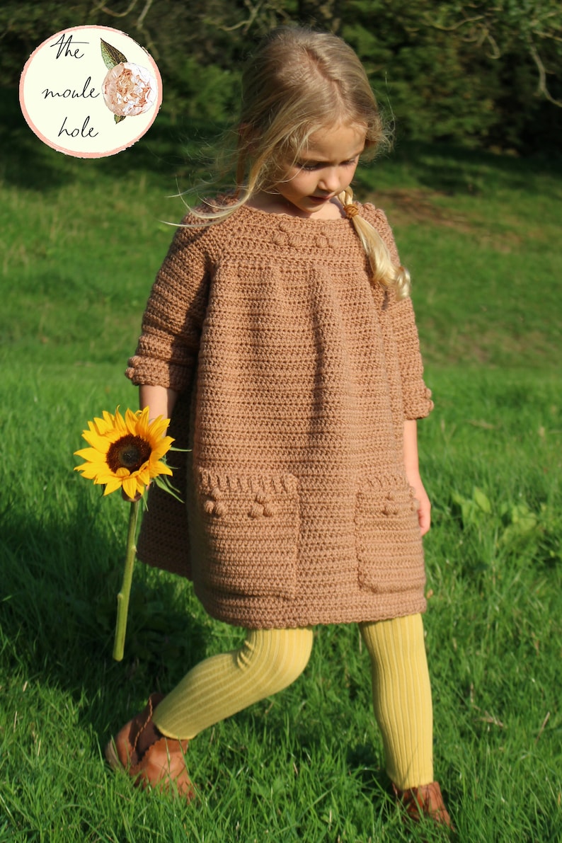 May include: A young girl wearing a brown crocheted dress with pockets and yellow tights. She is holding a sunflower and standing in a grassy field.
