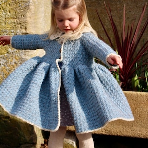 May include: A little girl wearing a light blue crocheted coat with a white crocheted collar and trim. The coat has a flared skirt and is worn over a patterned dress.