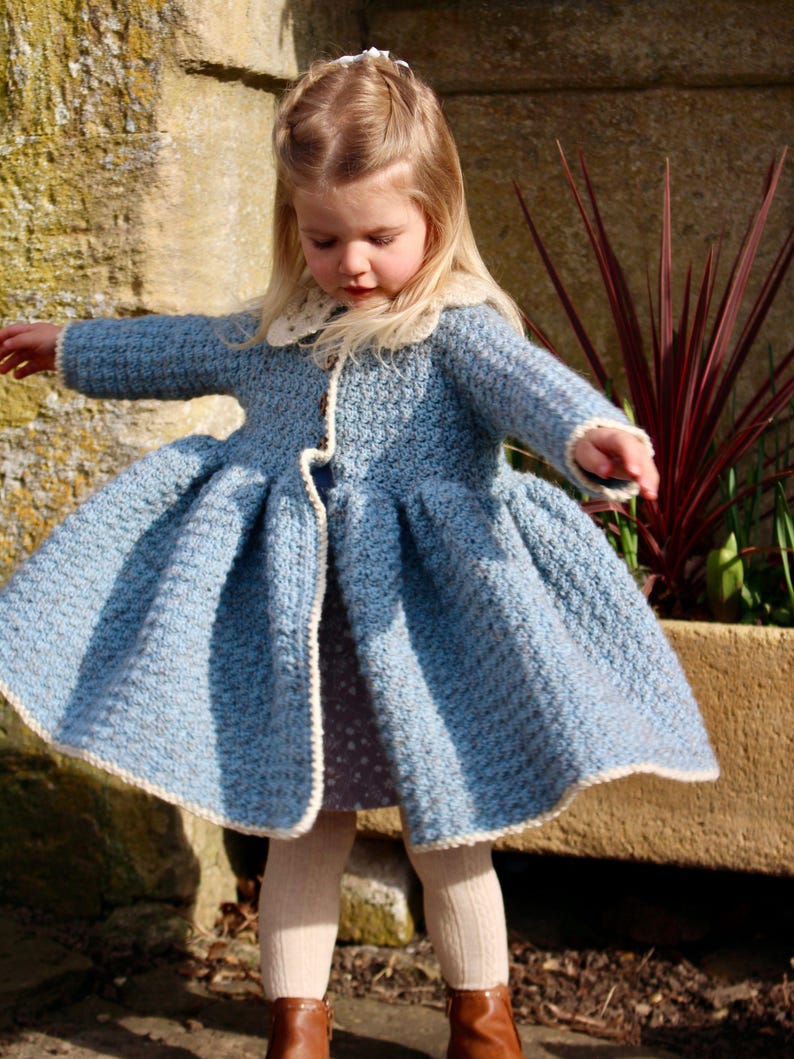 May include: A little girl wearing a light blue crocheted coat with a white crocheted collar and trim. The coat has a flared skirt and is worn over a patterned dress.