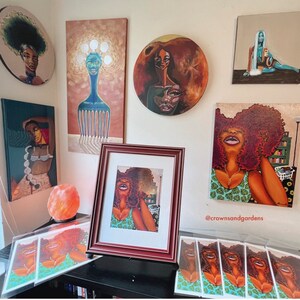 May include: A collection of framed artwork featuring colorful portraits of women with vibrant hair and clothing. The artwork is displayed on a wall with a salt lamp and a set of prints on a table below. The prints feature a woman with a green and white patterned dress and the text "@crownsandgardens".