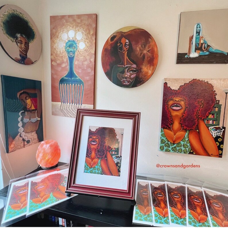May include: A collection of framed artwork featuring colorful portraits of women with vibrant hair and clothing. The artwork is displayed on a wall with a salt lamp and a set of prints on a table below. The prints feature a woman with a green and white patterned dress and the text "@crownsandgardens".