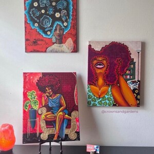 May include: Three paintings of women with afro hairstyles. The paintings are in a red, orange, and gold color scheme. The paintings are on a white wall with a black table in front of them. The table has a salt lamp and a business card on it.