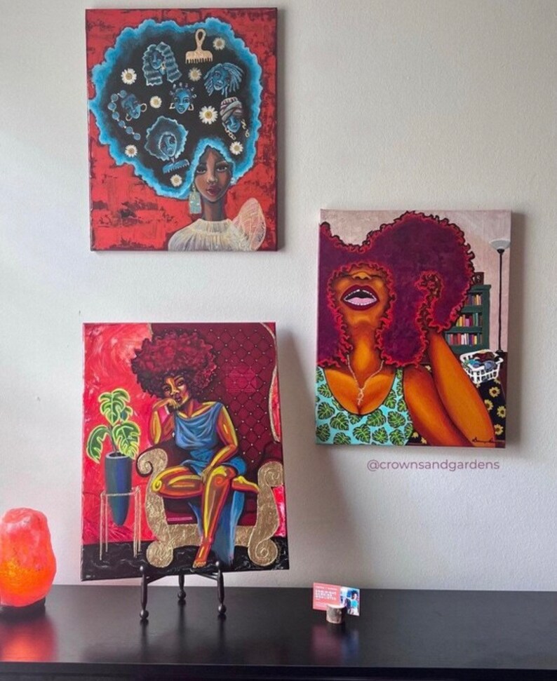 May include: Three paintings of women with afro hairstyles. The paintings are in a red, orange, and gold color scheme. The paintings are on a white wall with a black table in front of them. The table has a salt lamp and a business card on it.