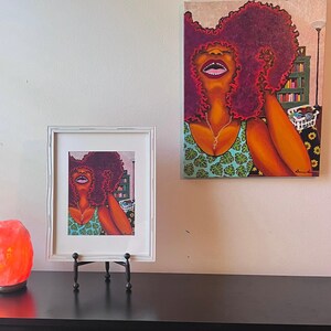 May include: Two framed paintings of a woman with red curly hair wearing a green and blue patterned top. The woman in the larger painting is looking up and smiling. The woman in the smaller painting is looking to the side. Both paintings have a white frame.