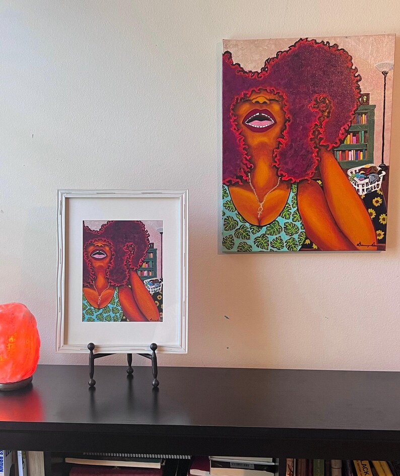 May include: Two framed paintings of a woman with red curly hair wearing a green and blue patterned top. The woman in the larger painting is looking up and smiling. The woman in the smaller painting is looking to the side. Both paintings have a white frame.