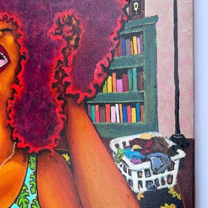 May include: A painting of a woman with a large, curly, red afro. She is wearing a blue and green tank top with a leafy pattern. The woman is sitting on a bed with a white laundry basket filled with clothes. A bookshelf with books is in the background.
