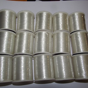 May include: A close-up of 15 spools of clear, white thread. The spools are arranged in three rows of five. The spools are all the same size and shape.