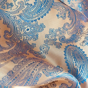 May include: A close-up of a fabric with a paisley pattern. The fabric is a light beige colour with blue paisley designs. The fabric is folded and draped.