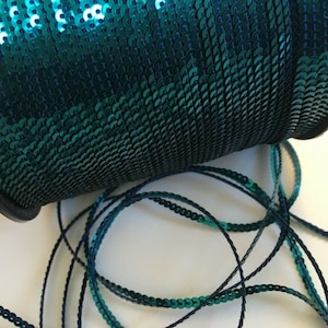May include: A spool of teal green sequined trim with a black thread running through the centre. The trim is rolled up on a black spool.