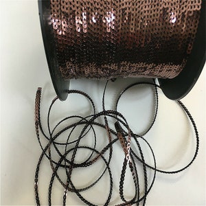 May include: A spool of brown sequined trim with a black thread running through the centre. The trim is coiled and ready to be used for crafting or decorating.