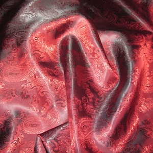 May include: Red paisley patterned fabric with a satin sheen. The fabric is draped and folded, showing the texture and pattern.