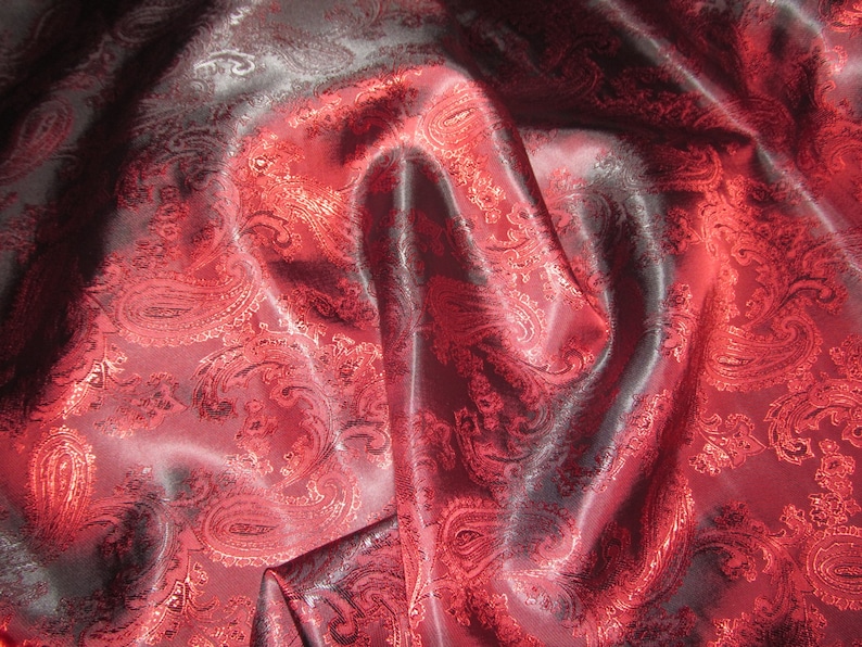 May include: Red paisley patterned fabric with a satin sheen. The fabric is draped and folded, showing the texture and pattern.