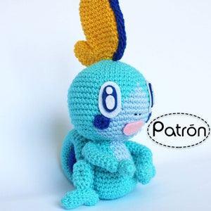 May include: A crocheted blue and yellow plush toy resembling a water-type Pokemon. The toy has a yellow fin on its head and a blue body with white eyes and a pink mouth. The toy is sitting on a white surface. The word "Patrón" is written in black on a white oval with a dashed border.