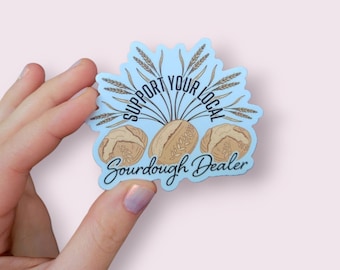 May include: A blue sticker with the text "Support Your Local Sourdough Dealer" in black lettering. The sticker features three loaves of sourdough bread and wheat stalks.