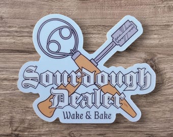 May include: A light blue sticker with the words "Sourdough Dealer" in a decorative font, along with "Wake & Bake". The sticker features illustrations of a dough whisk and a lame, both in shades of brown and gray.