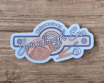 May include: A blue and white sticker with the text "Welcome to my Sourdough Era" in a retro font. The sticker features a loaf of sourdough bread, a bread lame, and a proofing basket.