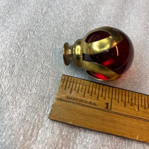 May include: A spherical red glass knob encased in a decorative brass cage with a metal mounting base. The knob is positioned next to a wooden ruler, showing the object is approximately 2.5 cm in diameter at its widest point, with the text MADE IN USA.