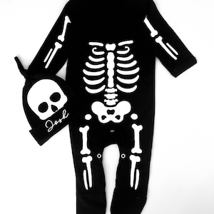 May include: Black skeleton onesie with a white skeleton design. The onesie has a matching skull hat with the name "Josh" written on it.