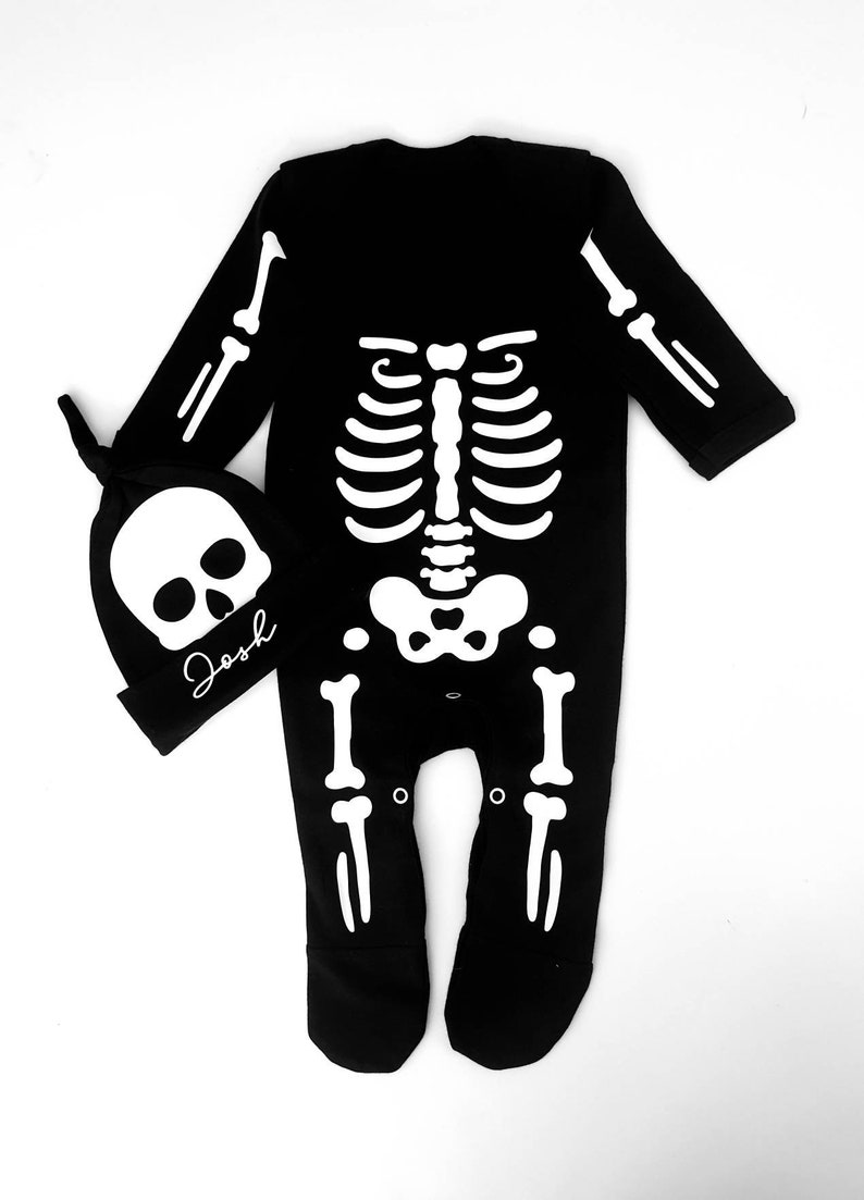May include: Black skeleton onesie with a white skeleton design. The onesie has a matching skull hat with the name "Josh" written on it.