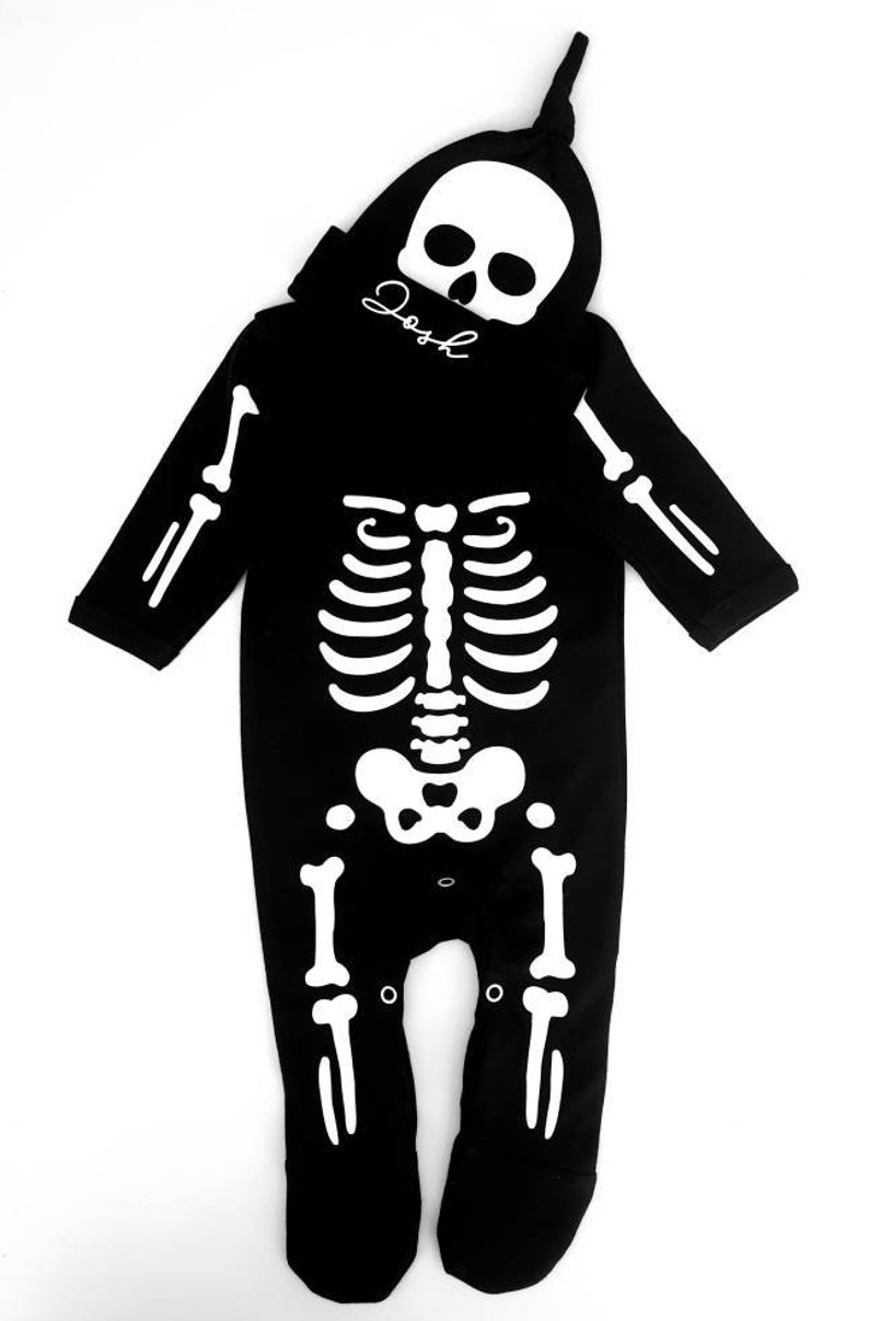 May include: Black skeleton costume with a white skeleton design. The costume includes a matching skull hat with the name "Josh" written on it.