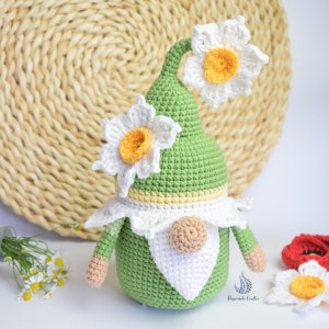 May include: A green crocheted gnome with a white beard and a white crocheted hat with two white flowers with yellow centres. The gnome is sitting on a woven straw mat.