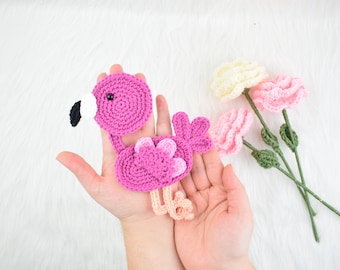 Puede incluir: Un flamenco de crochet rosa con detalles blancos y negros. El flamenco está siendo sostenido en las manos de una persona. Hay tres flores de crochet en el fondo.