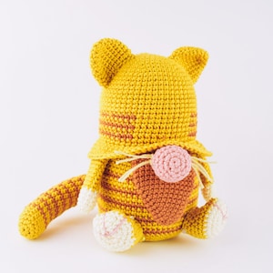 Peut inclure: Un jouet en peluche de chat crocheté avec des rayures jaunes et orange, un nez rose et des pattes blanches.