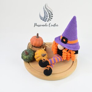 May include: A purple and orange crocheted gnome wearing a witch hat sits on a wooden board with three pumpkins. The gnome has a black beard and orange and white striped legs.