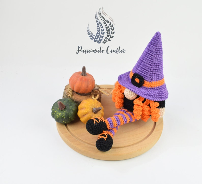 May include: A purple and orange crocheted gnome wearing a witch hat sits on a wooden board with three pumpkins. The gnome has a black beard and orange and white striped legs.