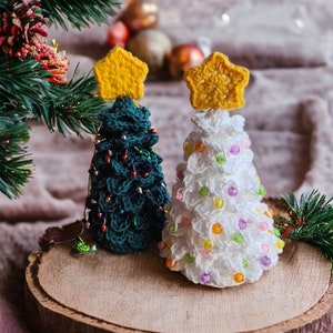 Può includere: Due alberi di Natale all'uncinetto, uno verde e uno bianco, con stelle gialle in cima e decorazioni di perline colorate, su una fetta di legno.