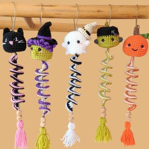 May include: Five hanging crochet ornaments featuring a black cat, a witch, a ghost, a Frankenstein-style monster, and a pumpkin. Each figure has a spiral tail, wooden beads, and a colorful yarn tassel, all suspended from silver metal hooks.