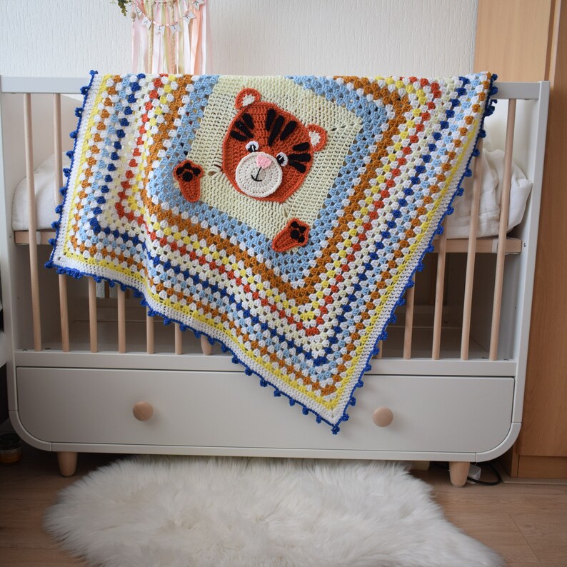 Peut inclure: Une couverture de b&eacute;b&eacute; en crochet color&eacute;e avec une applique de tigre au centre. La couverture est faite de plusieurs couleurs, dont le bleu, le jaune, l'orange et le blanc. La couverture est drap&eacute;e sur un lit b&eacute;b&eacute; blanc avec un cadre en bois.