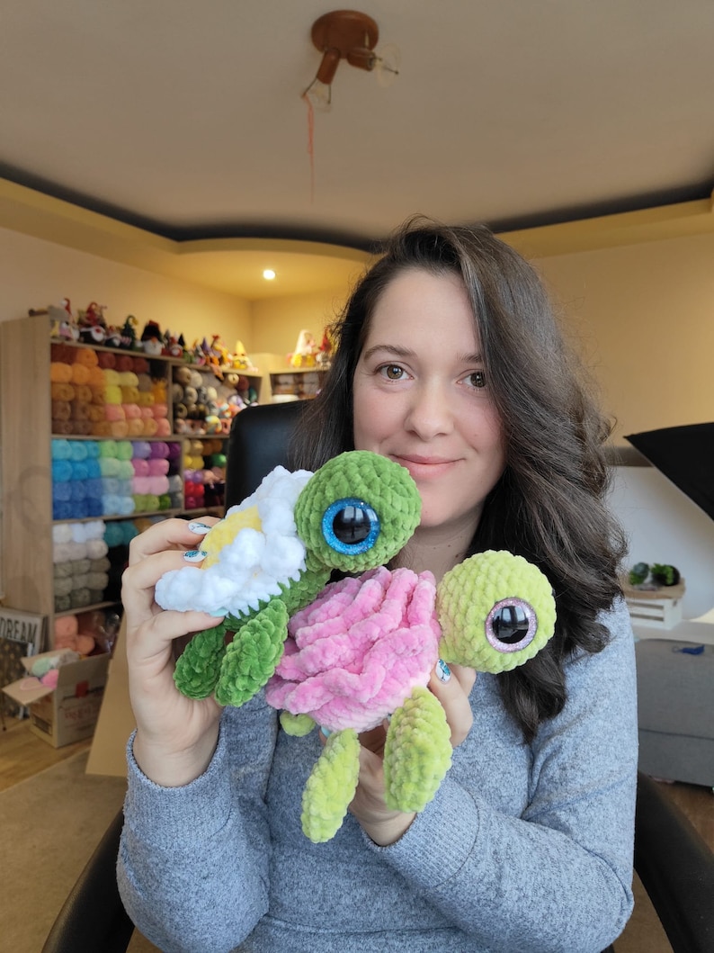 May include: Two crocheted turtle plushies, one green with a white flower and one pink, held by a person. The turtles have large, sparkly eyes.
